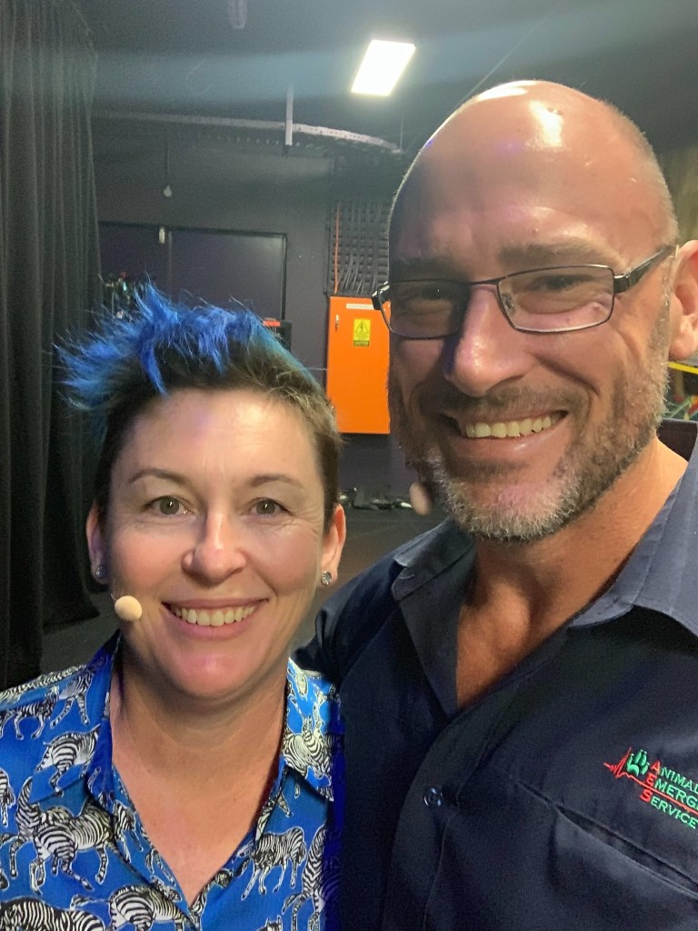 Dr Jodie stands backstage. She is wearing a headset microphone and a blue button down shirt covered in black and white zebras. She stands next to a man who has his arms over her shoulder. Both people are smiling at the camera.