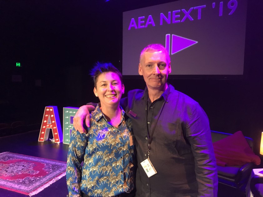 Dr Jodie stands in front of a stage. She is wearing a headset microphone and a blue button down shirt covered in black and white zebras. She stands next to a man who has his arms over her shoulder. Both people are smiling at the camera.